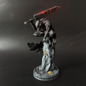 Puede incluir: Una figura en miniatura detallada de un guerrero con armadura oscura y texturizada sobre una base rocosa. El personaje sostiene una espada grande y pesada sobre los hombros, con salpicaduras de pintura roja en la hoja y una capa negra.