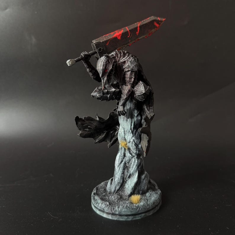 Puede incluir: Una figura en miniatura detallada de un guerrero con armadura oscura y texturizada sobre una base rocosa. El personaje sostiene una espada grande y pesada sobre los hombros, con salpicaduras de pintura roja en la hoja y una capa negra.