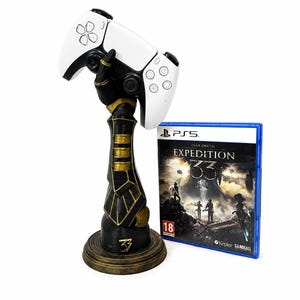 May include: A black and gold gauntlet-shaped controller stand holding a white wireless game controller, positioned next to a blue-cased video game for PS5 titled Clair Obscur Expedition 33 with an 18 age rating symbol on the front cover.