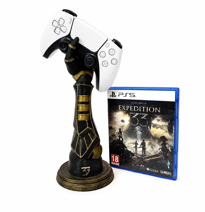 May include: A black and gold gauntlet-shaped controller stand holding a white wireless game controller, positioned next to a blue-cased video game for PS5 titled Clair Obscur Expedition 33 with an 18 age rating symbol on the front cover.