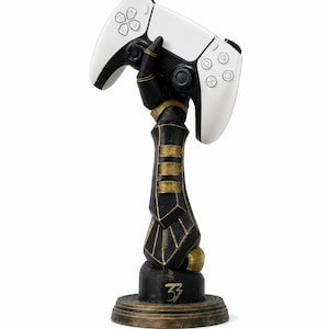May include: A black and gold robotic arm sculpture mounted on a circular base, featuring the number 33 in gold on the front. The hand of the sculpture is positioned to hold a white and black wireless video game controller with standard buttons.