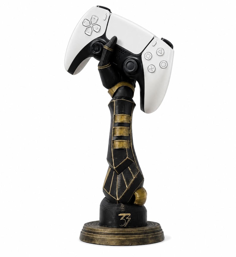 May include: A black and gold robotic arm sculpture mounted on a circular base, featuring the number 33 in gold on the front. The hand of the sculpture is positioned to hold a white and black wireless video game controller with standard buttons.