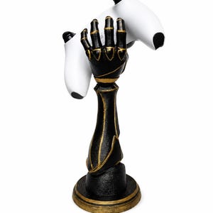 May include: A black and gold robotic hand sculpture mounted on a circular base, designed to hold a white video game controller. The hand features articulated fingers and metallic gold accents along the joints and forearm, providing a stable stand.