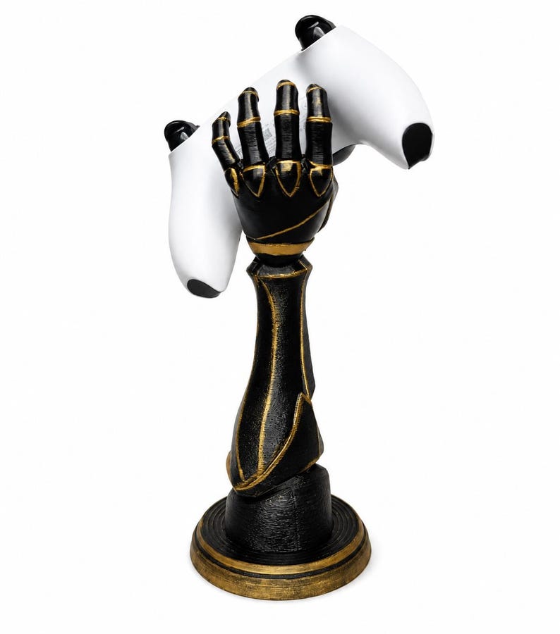May include: A black and gold robotic hand sculpture mounted on a circular base, designed to hold a white video game controller. The hand features articulated fingers and metallic gold accents along the joints and forearm, providing a stable stand.