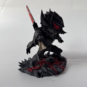 Puede incluir: Una figura negra de un guerrero en una pose din&aacute;mica, empu&ntilde;ando una gran espada con una hoja roja. El guerrero est&aacute; vestido con una armadura detallada, con detalles rojos en el casco. La base presenta una superficie oscura y texturizada con elementos rojos.