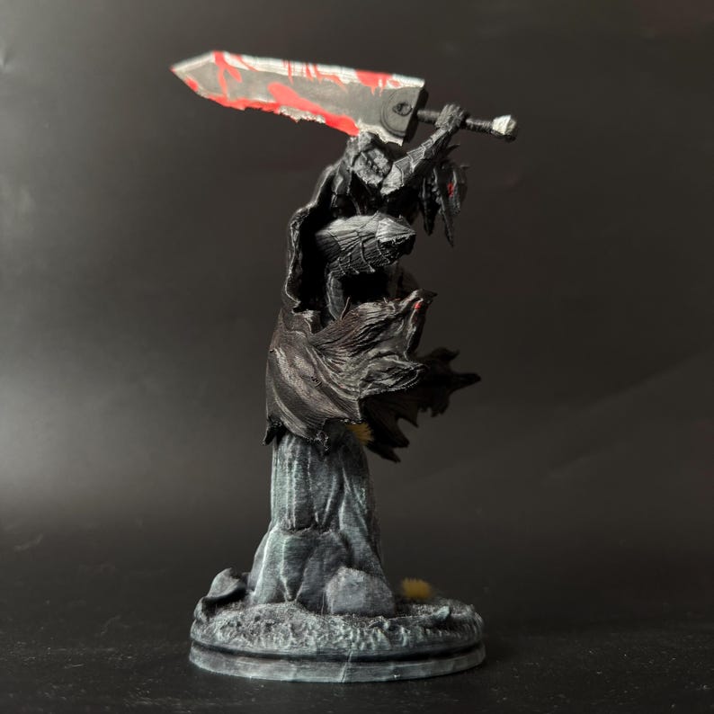 Puede incluir: Una figura pintada de un guerrero con armadura negra y capa ondeante, sobre una base gris texturizada. El personaje sostiene una espada grande y pesada sobre el hombro, con una hoja met&aacute;lica que presenta salpicaduras de pintura roja.