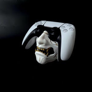 May include: A white and black PlayStation 5 controller frames a white and gold Oni mask. The mask has a detailed, textured surface with sharp teeth. The background is solid black.