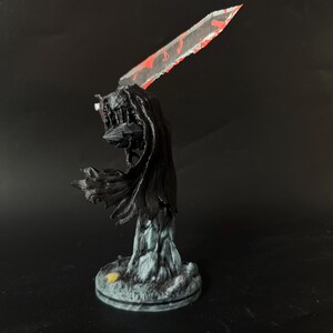 Puede incluir: Una figura en miniatura de un guerrero con una capa negra fluida y armadura, sosteniendo una gran espada transl&uacute;cida con detalles pintados en rojo. La figura se apoya sobre una base texturizada de color gris claro sobre fondo negro.