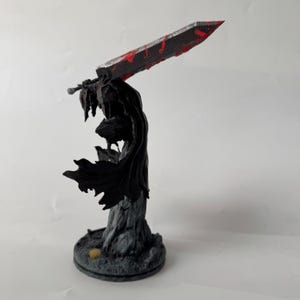 Puede incluir: Una figura detallada de un guerrero con capa sobre una base texturizada, sosteniendo una espada grande y pesada sobre el hombro. La espada presenta una hoja oscura y desgastada con salpicaduras de pintura roja brillante, y el guerrero lleva una capa negra.