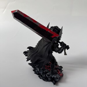 Puede incluir: Figura negra de un guerrero sosteniendo una gran espada con detalles rojos. El guerrero lleva una armadura y una capa, de pie sobre una base texturizada con detalles rojos. La espada tiene un dise&ntilde;o sangriento.