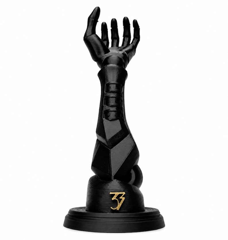 May include: A matte black sculptural object depicting a robotic arm extending upwards with an open hand. The forearm features segmented geometric plating. At the base, a circular stand displays the gold-coloured number 33 in a stylised font.
