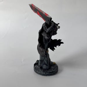 Puede incluir: Una figura detallada de un guerrero con armadura oscura y una capa negra que fluye, sobre una base circular texturizada. El personaje sostiene una espada masiva sobre el hombro, con una hoja oscura que presenta salpicaduras de color rojo.