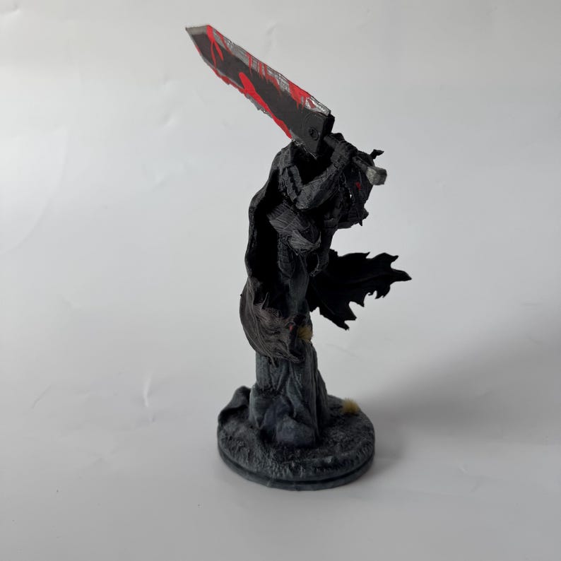 Puede incluir: Una figura detallada de un guerrero con armadura oscura y una capa negra que fluye, sobre una base circular texturizada. El personaje sostiene una espada masiva sobre el hombro, con una hoja oscura que presenta salpicaduras de color rojo.