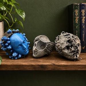 Puede incluir: Tres esculturas decorativas con rostros humanos. La escultura de la izquierda es de color azul brillante con protuberancias cristalinas. La central es gris con un detalle de ojo en espiral. La de la derecha es gris con una superficie texturizada.