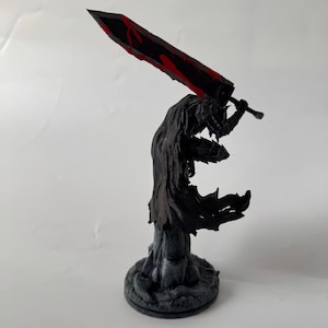 Puede incluir: Una figura detallada de un guerrero con armadura negra y capa fluida, sobre una base texturizada. El personaje sostiene una espada masiva sobre el hombro, con una hoja negra y llamativos detalles en rojo a lo largo del filo.