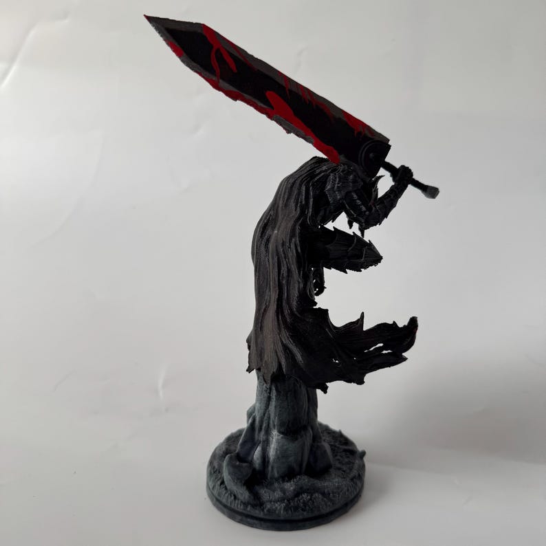 Puede incluir: Una figura detallada de un guerrero con armadura negra y capa fluida, sobre una base texturizada. El personaje sostiene una espada masiva sobre el hombro, con una hoja negra y llamativos detalles en rojo a lo largo del filo.