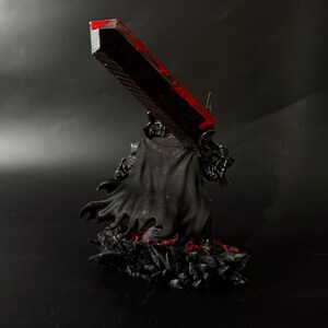 Puede incluir: Una figura negra de un personaje de la serie de manga Berserk, sosteniendo una gran espada con una hoja roja. La figura lleva una capa y armadura negras, de pie sobre una base con detalles rojos.