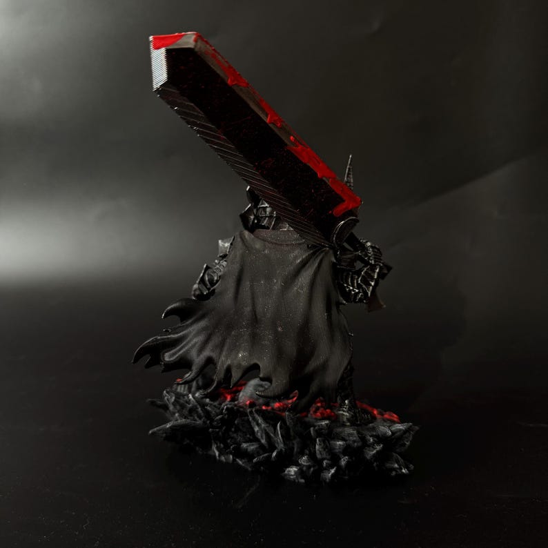 Puede incluir: Una figura negra de un personaje de la serie de manga Berserk, sosteniendo una gran espada con una hoja roja. La figura lleva una capa y armadura negras, de pie sobre una base con detalles rojos.