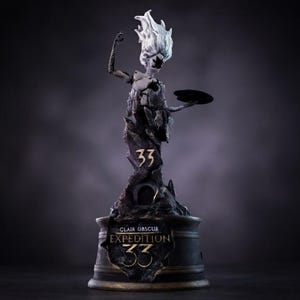 Könnte beinhalten: Dunkle Statue einer Figur mit fließendem weißem Kopf, die eine Scheibe hält. Die Statue trägt den Text "Clair Obscur Expedition 33" in goldfarbenen Buchstaben. Die Statue steht auf einem dunklen Sockel.