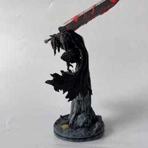 Puede incluir: Una figura en miniatura pintada de un guerrero con una capa negra fluida y armadura, sobre una base rocosa. La figura sostiene una espada grande y pesada en la espalda, con tonos gris met&aacute;lico y salpicaduras de pintura roja brillante.