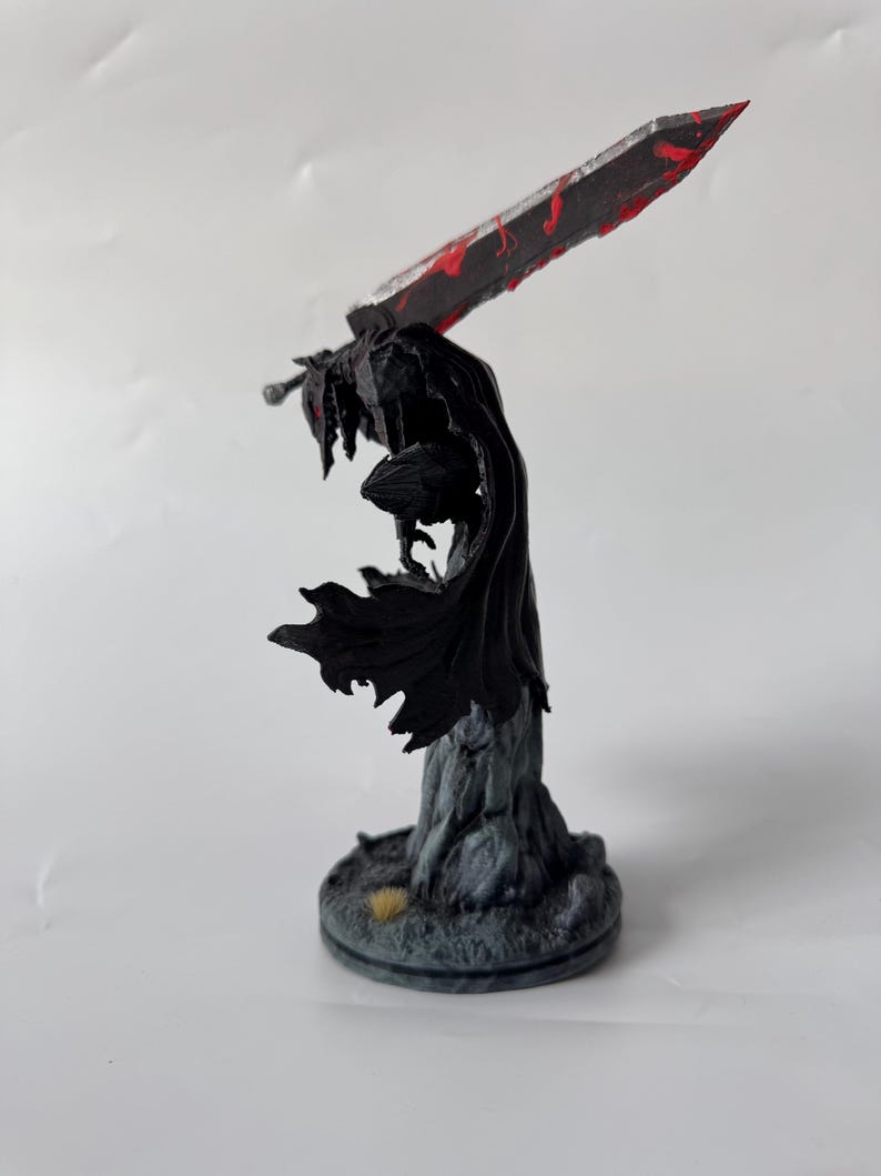 Puede incluir: Una figura en miniatura pintada de un guerrero con una capa negra fluida y armadura, sobre una base rocosa. La figura sostiene una espada grande y pesada en la espalda, con tonos gris met&aacute;lico y salpicaduras de pintura roja brillante.