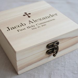 May include: A light-colored wooden box with a hinged lid and a metal clasp. The box is engraved with a cross and the name "Jacob Alexander", along with "First Holy Communion" and the date "May 20, 2026".