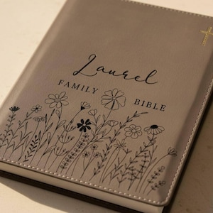 May include: A taupe-colored Bible with the name "Laurel" and the words "Family Bible" printed on the cover. The cover features a floral design and a gold cross. The edges of the Bible are brown.