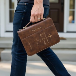 May include: A brown leather Bible cover with a handle, featuring the name "Liam Powell" and the inscription "Let all that you do be done with Love" along with a cross. The cover is held by a person wearing blue jeans.
