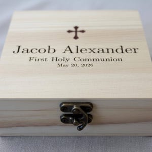 May include: A light-colored wooden box with a cross symbol and the inscription "Jacob Alexander First Holy Communion May 20, 2026." The box has a metal clasp and is likely used for religious keepsakes.