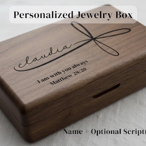 May include: A rectangular wooden jewelry box with a dark brown finish. The box is personalized with the name "claudia" and a cross design. The inscription "I am with you always Matthew 28:20" is also visible. The text "Name + Optional Scripture" is at the bottom.