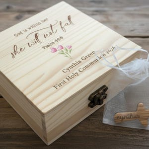 May include: A light-colored wooden box with a hinged lid, featuring engraved text and floral accents. The box includes the inscription "God is within her, she will not fall, Psalm 46:5," along with a name and date. A small wooden cross with a name is in a sheer bag.