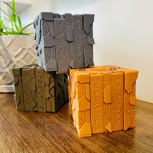 May include: Three 3D-printed puzzle cubes in gray, olive green, and orange. Each cube has a unique design with engraved text. The cubes are stacked on a wooden surface, with a white patterned pot and plant in the background.