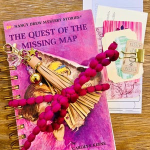 May include: A pink spiral-bound notebook with gold accents and the title "The Quest of the Missing Map" by Carolyn Keene. A fuchsia beaded dragonfly and a gold charm with a star and tassel rest on the book. Assorted vintage ephemera is visible.