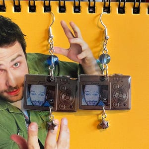 May include: A pair of silver-toned earrings featuring miniature camera pendants. Each camera pendant displays a small image of a person's face on the screen. The earrings have blue bead accents and star charms dangling below the cameras. The word "Canon" is visible on the camera.