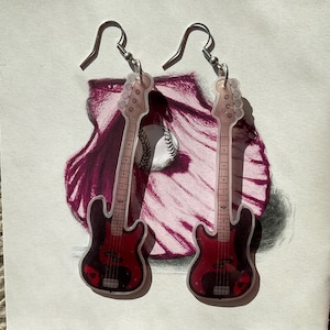 Puede incluir: Un par de pendientes con forma de guitarra con ganchos plateados. Los pendientes son rojos y negros, con una pelota de béisbol y un dibujo granate en el fondo. Los pendientes son un accesorio único para los amantes de la música.