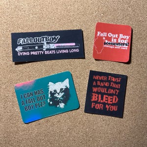 Op de afbeelding: Vier rechthoekige en vierkante stickers met tekst en afbeeldingen. Eén zwarte sticker met "FALL OUT BOY" en "DYING PRETTY BEATS LIVING LONG". Een rode sticker zegt "Fall Out Boy is for scenesters!" met een Illinois-omtrek. Een blauwgroene sticker heeft een kattenafbeelding en "I CAN HAS A FALL OUT BOY PLZ?". Een zwarte sticker zegt "NEVER TRUST A BAND THAT WOULDN'T BLEED FOR YOU".