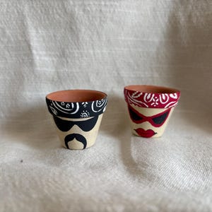 May include: Two small terracotta pots, hand-painted with faces. One pot features a black bandana, sunglasses, and a mustache. The other pot has a red bandana, sunglasses, and red lips. The pots are on a neutral background.