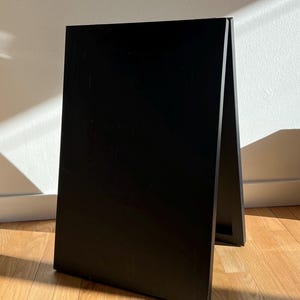 May include: A black, freestanding chalkboard sign with a simple A-frame design. The sign is made of wood and stands on a hardwood floor. The background is a white wall with natural light.