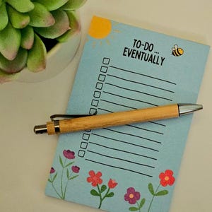 May include: A pale blue notepad with the text "TO-DO... EVENTUALLY" and a sun and bee illustration. A wooden pen rests on the notepad, which has a floral design at the bottom. A succulent plant is in the background.
