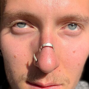 May include: Close-up of a person's face with blue eyes and a silver nose ring. The nose ring is a curved design that wraps around the septum. A second silver ring is in the nostril.