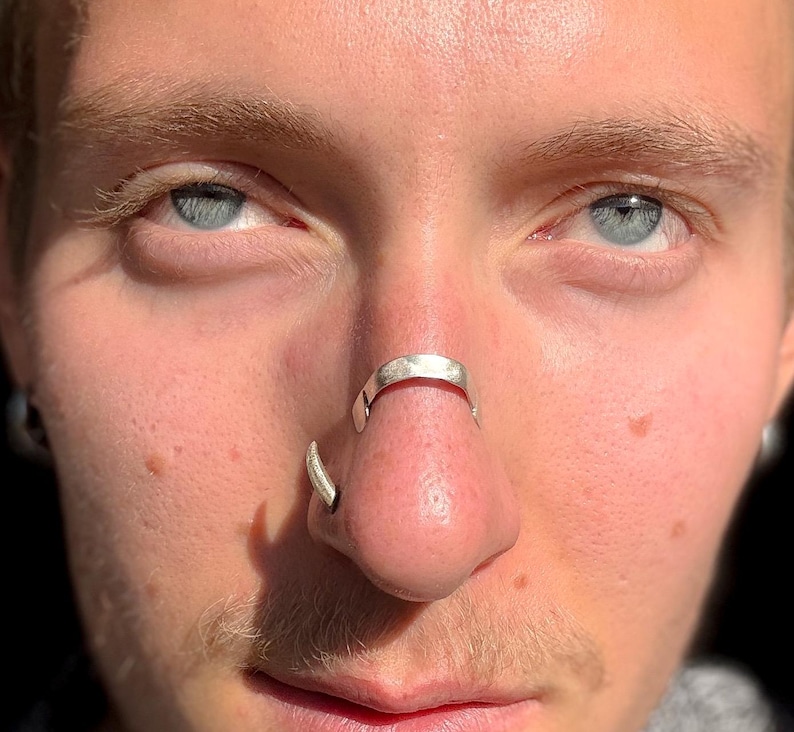 May include: Close-up of a person's face with blue eyes and a silver nose ring. The nose ring is a curved design that wraps around the septum. A second silver ring is in the nostril.