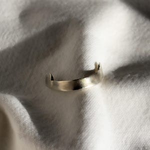 May include: A minimalist silver ring with a brushed finish. The ring has a curved band with two small, raised points on either side. The ring is set against a white, textured background, creating a contrast of light and shadow.
