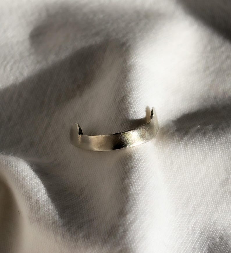 May include: A minimalist silver ring with a brushed finish. The ring has a curved band with two small, raised points on either side. The ring is set against a white, textured background, creating a contrast of light and shadow.