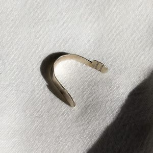 May include: A silver-colored, curved metal object, possibly a bracelet or cuff, lies on a white, textured surface. The object has a smooth, polished finish and a simple, minimalist design. The shape is reminiscent of a partial arc, with a small, rectangular detail at one end.