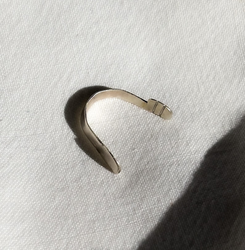 May include: A silver-colored, curved metal object, possibly a bracelet or cuff, lies on a white, textured surface. The object has a smooth, polished finish and a simple, minimalist design. The shape is reminiscent of a partial arc, with a small, rectangular detail at one end.