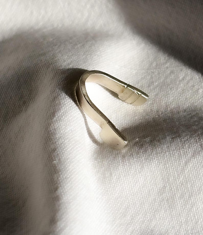 May include: A silver-colored, curved metal object, possibly a jewelry clasp or finding, rests on a white, textured fabric. The object has a smooth, reflective surface and a simple, minimalist design. The lighting creates shadows that highlight its shape.