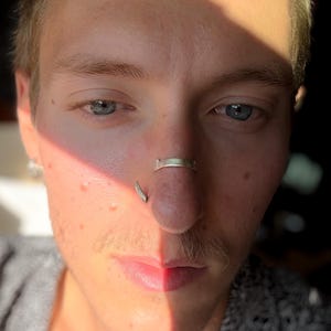 May include: Close-up of a person's face with blue eyes, a mustache, and two nose piercings. One piercing is a silver ring on the septum, and the other is a curved silver stud on the nostril. The face is partially lit by sunlight.