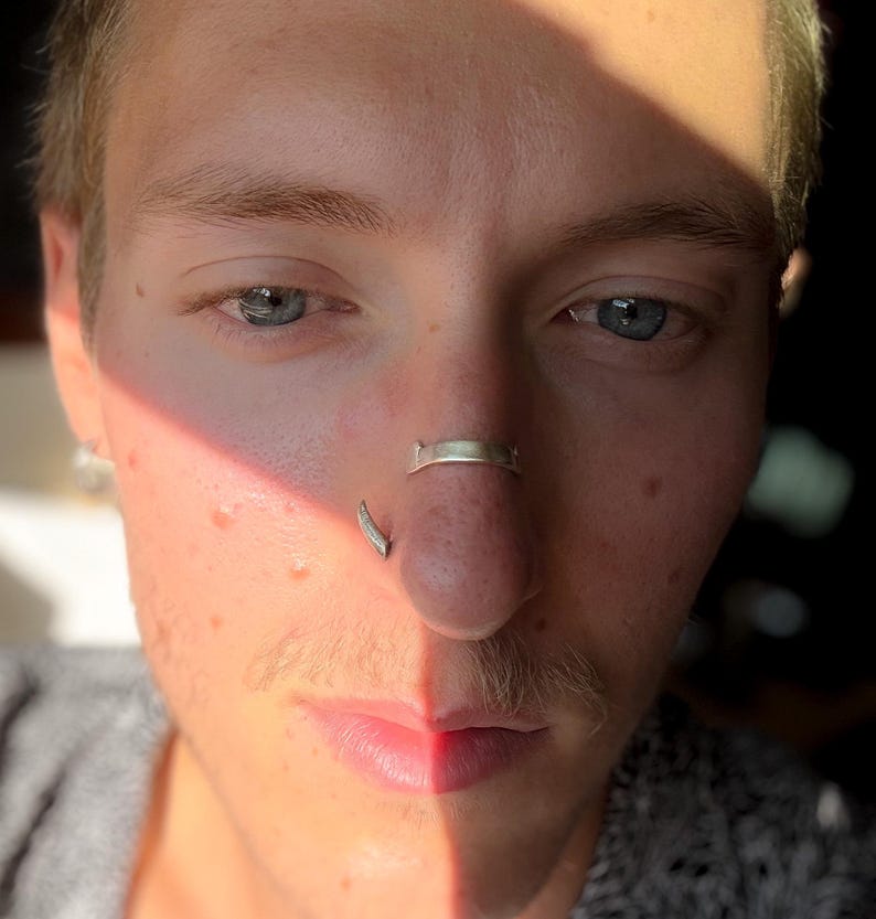 May include: Close-up of a person's face with blue eyes, a mustache, and two nose piercings. One piercing is a silver ring on the septum, and the other is a curved silver stud on the nostril. The face is partially lit by sunlight.