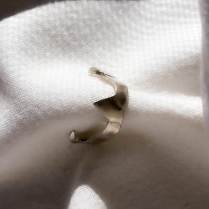 May include: A silver-colored ring with a unique, abstract design. The ring features a curved shape with a pointed element, creating a modern and artistic look. The ring is set against a white background.