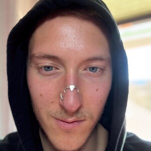 May include: A silver-colored nose ring with a geometric design. The nose ring has a triangular center piece and curved side elements. The person is wearing a black hooded sweatshirt.